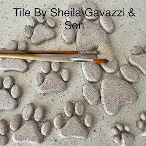 May include: A collection of gray ceramic paw print tiles with a speckled finish. The tiles are arranged in a semi-circle pattern on a light gray surface. Two paintbrushes are placed on top of the tiles. The text "Tile By Sheila Gavazzi & Son" is visible at the top of the image.