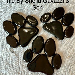 May include: Arrangement of dark brown, glossy tiles in the shape of a paw print, with the text "Tile By Sheila Gavazzi & Son" at the top. The tiles are oval and irregularly shaped.