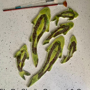 May include: A set of six green and brown ceramic fish tiles, arranged in a decorative pattern. The tiles are hand-painted with a mottled glaze and have a slightly textured surface. The tiles are arranged in a way that suggests they are swimming in a school. The text "Tile By Sheila Gavazzi & Son" is visible at the bottom of the image.