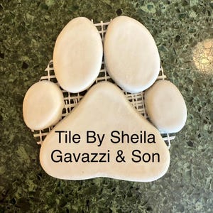 May include: A paw-shaped tile mosaic made of white stones, set on a white grid. The tile has the text "Tile By Sheila Gavazzi & Son" in black. The background is a dark green, speckled surface.