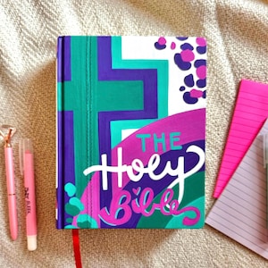 May include: A hardcover book titled "The Holy Bible" with a colorful design. The cover features a teal cross, purple and teal geometric shapes, and a pink and purple design with the title in white script. Pink pens and a notepad are next to the book.