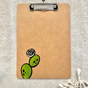 May include: A brown clipboard with a silver clip and a hand-painted green cactus with a white flower on the bottom right corner.