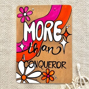 May include: A brown cardboard sign with a pink and orange abstract design. The sign says "MORE than A CONQUEROR" in white lettering.