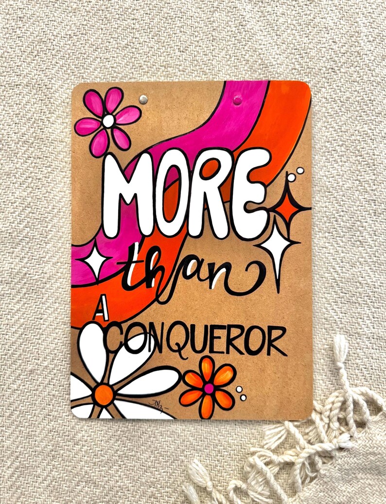 May include: A brown cardboard sign with a pink and orange abstract design. The sign says "MORE than A CONQUEROR" in white lettering.