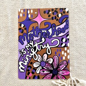 May include: Purple and brown background with a pink and white flower. The text "Motherhood is my ministry" is written in a purple, white, and black font.