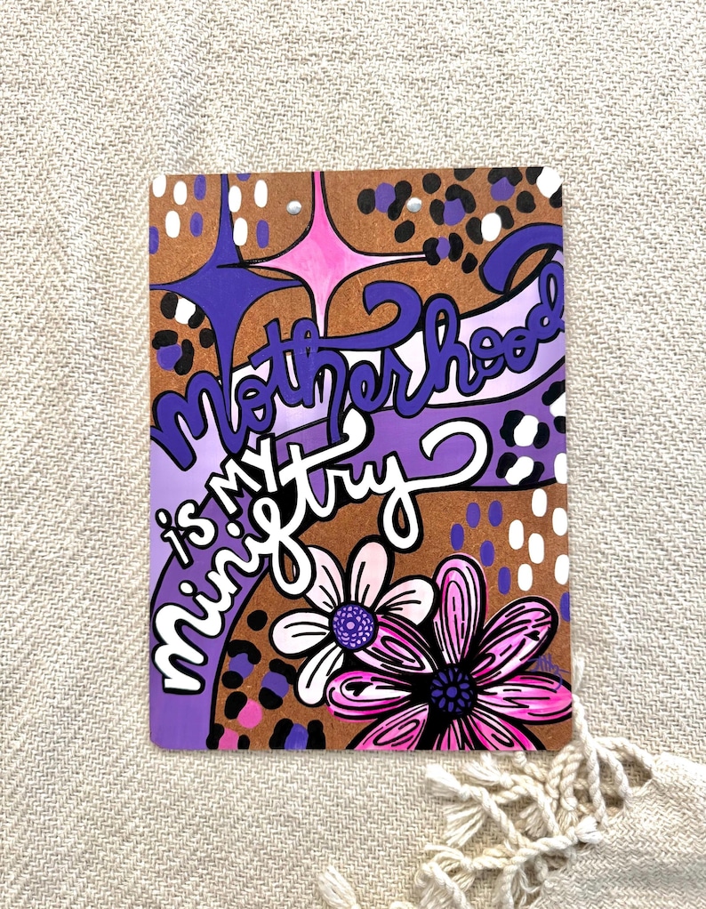 May include: Purple and brown background with a pink and white flower. The text "Motherhood is my ministry" is written in a purple, white, and black font.