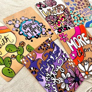 May include: Six different clipboard signs with inspirational quotes and designs. The signs feature a leopard print background with flowers, cacti, and other designs. The quotes include "Blessed Mama", "WWID", "More Than A Conquer", "Created For A Purpose", "With Faith", and "Motherhood is my greatest adventure".