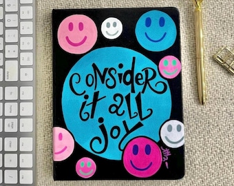 May include: A black notebook with a blue circle and the text "Consider it all joy" in white. The circle is surrounded by colorful smiley faces.