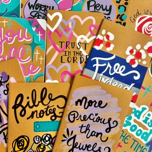 May include: A collection of brown notebooks with painted designs and inspirational quotes. The quotes include "Trust in the Lord", "Free Indeed", "More Precious than Jewels", "Bible Notes", and "God is with you".