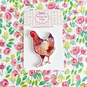May include: An embroidered hen patch in shades of red, brown, and cream, displayed on a white card. The card features the text "Makes by Kelly etsy.com" and is set against a floral pattern with pink flowers.