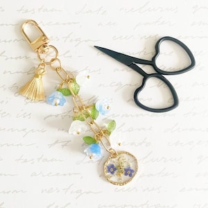 May include: A gold-toned keyring featuring a lobster clasp, a small gold tassel, and a chain adorned with translucent blue and white bell-shaped flower charms, green leaves, and a circular resin pendant containing pressed blue flowers.