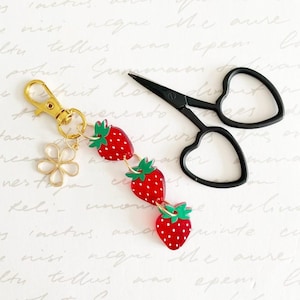 May include: A gold-toned metal keyring featuring three red strawberry charms with green leaves and white dots, accompanied by a small gold flower charm. Next to it lies a pair of black embroidery scissors with heart-shaped finger loops.