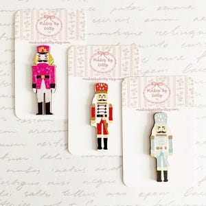 May include: Three wooden nutcracker pins displayed on individual white backing cards labelled Makes By Kelly. The pins feature a bright pink, a traditional red, and a light blue nutcracker design, each with black boots and detailed uniform accents.