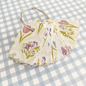 May include: A collection of floral-patterned index cards held together by a silver ring. The cards display a variety of flowers in purple, pink, and yellow hues on a white backdrop. The cards are fanned out, revealing the different floral patterns.