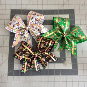 May include: Three Mardi Gras themed hair bows with gold glitter accents. The bows are made of black, white, and green ribbon with a Mardi Gras mask pattern.
