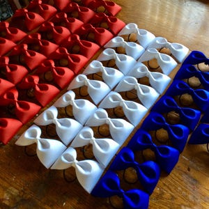 May include: A collection of red, white, and blue fabric bows arranged in rows. The bows are tied with elastic bands and are ready to be used as hair accessories.