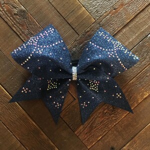 May include: A navy blue cheer bow with iridescent rhinestones. The bow is made of glitter fabric and has a large, full shape.