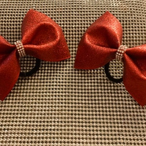 May include: Two red glitter hair bows with clear rhinestone centers. The bows are on a silver background.