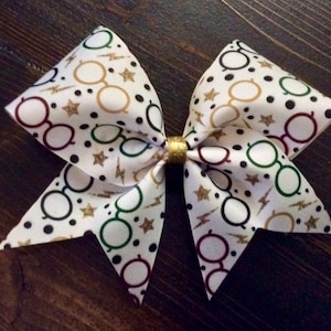 May include: A white cheer bow with a Harry Potter themed print. The print features glasses, stars, and lightning bolts in gold, black, green, and maroon.