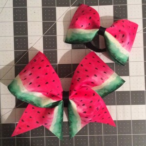 May include: Two watermelon-patterned hair bows, one large and one small, with black elastic bands. The bows are on a gray cutting mat.