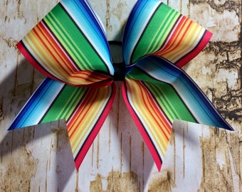 May include: A large, colorful bow with stripes of red, orange, yellow, green, blue, and white. The bow is made of a thick, woven fabric and is tied in a classic bow shape.