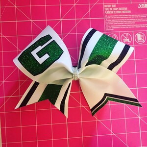 May include: A white cheer bow with green and black stripes. The bow has a large green glitter letter "G" on one side.