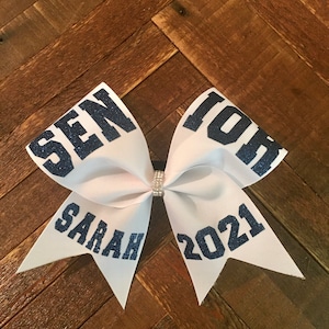 May include: White cheer bow with blue glitter lettering that says "Senior 2021" and "Sarah".