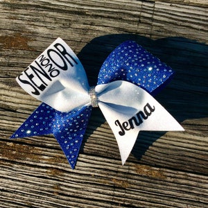 May include: A white and royal blue cheer bow with rhinestones. The bow has the text "Senior 2020" on one side and the name "Jenna" on the other side.