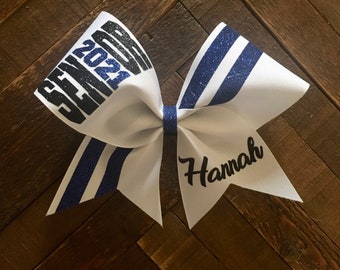May include: A white and blue graduation bow with the text "SENIOR 2021" in black glitter. The bow has blue glitter stripes and the name "Hannah" in black script. The bow is on a wooden surface.