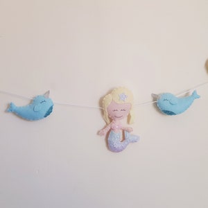 May include: A felt garland with three mermaids and two narwhals. The mermaids have blonde hair and pink and purple tails. The narwhals are blue. The garland is strung on a white string.