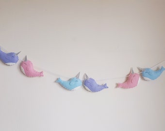 May include: A garland of felt narwhals in pink, blue, and purple with silver horns. The narwhals are strung on a white string.