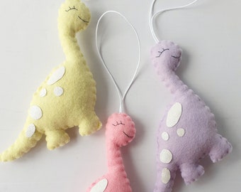 May include: Three felt dinosaur ornaments in yellow, purple, and pink with white spots and smiling faces. Each dinosaur has a white string loop for hanging.