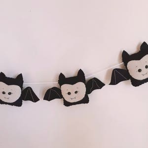 May include: A Halloween banner with three plush bat decorations. Each bat has a black body, wings, and ears, with a light grey face and small black eyes. The bats are strung on a white string, ideal for seasonal decorating.