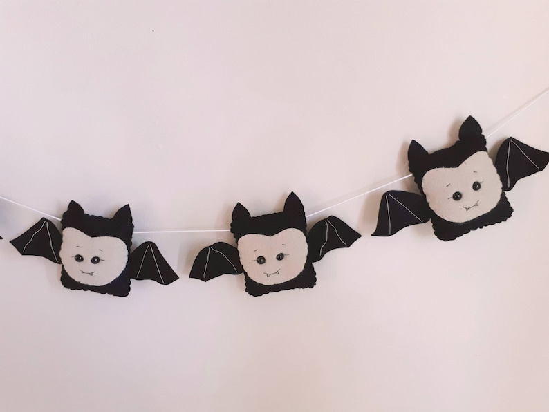 May include: A Halloween banner with three plush bat decorations. Each bat has a black body, wings, and ears, with a light grey face and small black eyes. The bats are strung on a white string, ideal for seasonal decorating.