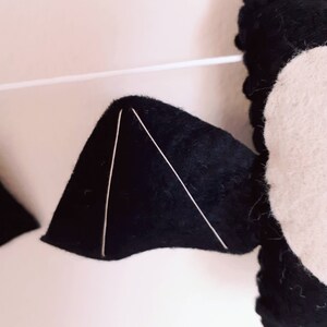 May include: Close-up of a felt bat decoration with black wings and a white body. The wings have white stitching to define the shape. The bat is strung on a white string, suggesting a garland or hanging decoration.