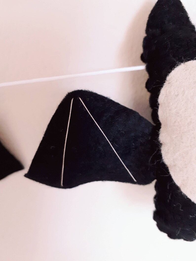May include: Close-up of a felt bat decoration with black wings and a white body. The wings have white stitching to define the shape. The bat is strung on a white string, suggesting a garland or hanging decoration.