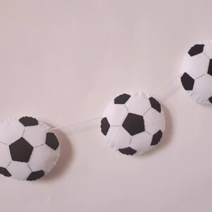 Puede incluir: Una guirnalda de bolas de f&uacute;tbol de fieltro blanco y negro con cinco bolas de f&uacute;tbol colgando de una cuerda blanca.