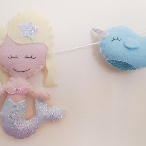 May include: A felt garland with a mermaid and a narwhal. The mermaid has blonde hair, a pink top, and a glittery tail. The narwhal is blue with a white horn.