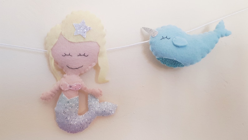 May include: A felt garland with a mermaid and a narwhal. The mermaid has blonde hair, a pink top, and a glittery tail. The narwhal is blue with a white horn.