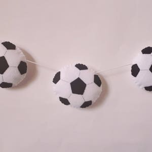 Puede incluir: Una guirnalda de fieltro blanco con balones de f&uacute;tbol de fieltro negro. La guirnalda est&aacute; enhebrada con hilo blanco.