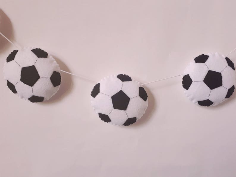 Puede incluir: Una guirnalda de fieltro blanco con balones de f&uacute;tbol de fieltro negro. La guirnalda est&aacute; enhebrada con hilo blanco.