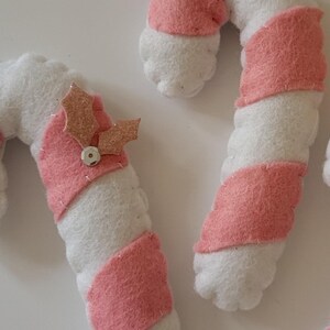 May include: Close-up of handmade Christmas ornaments shaped like candy canes. The candy canes are white with pink stripes and are topped with a pink glittery holly leaf and a silver bead. These festive decorations are perfect for the holiday season.
