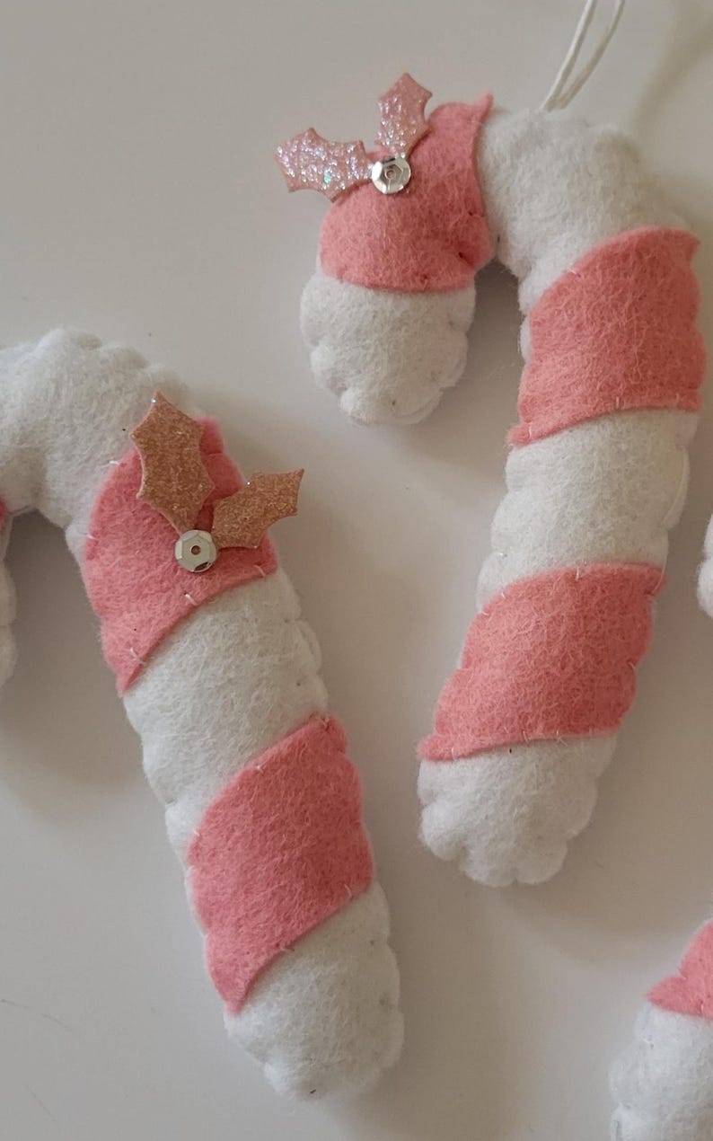 May include: Close-up of handmade Christmas ornaments shaped like candy canes. The candy canes are white with pink stripes and are topped with a pink glittery holly leaf and a silver bead. These festive decorations are perfect for the holiday season.