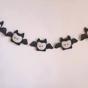 May include: A Halloween garland featuring five plush bat decorations. Each bat has a black body, grey wings, and a cream-coloured face with embroidered details. The bats are strung on a white cord.