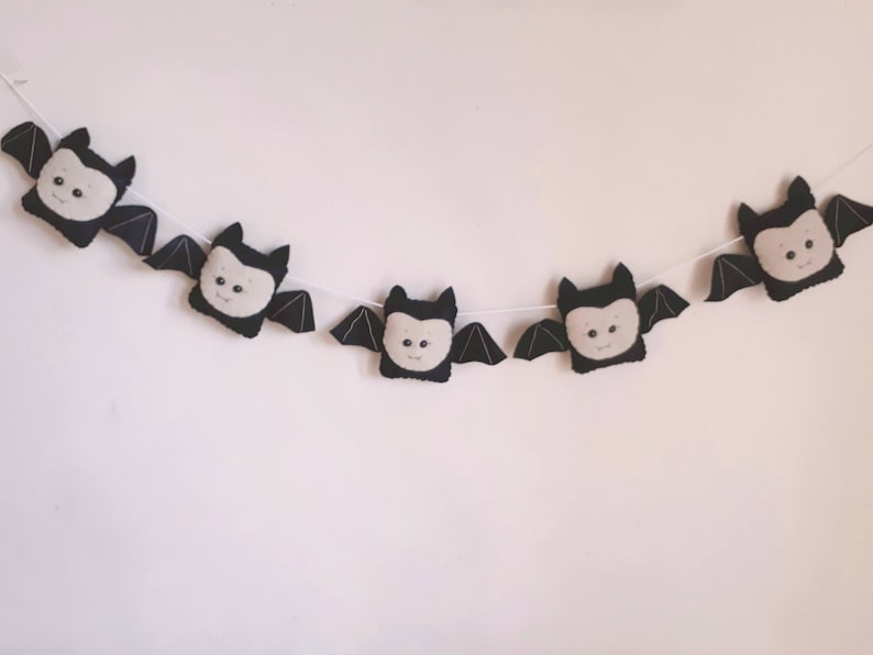 May include: A Halloween garland featuring five plush bat decorations. Each bat has a black body, grey wings, and a cream-coloured face with embroidered details. The bats are strung on a white cord.