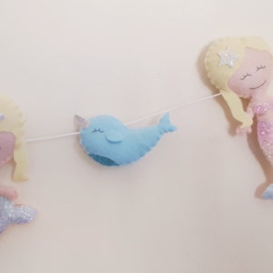 May include: A felt garland with two mermaids and a narwhal. The mermaids have blonde hair and pink and blue glitter tails. The narwhal is blue with a white horn.