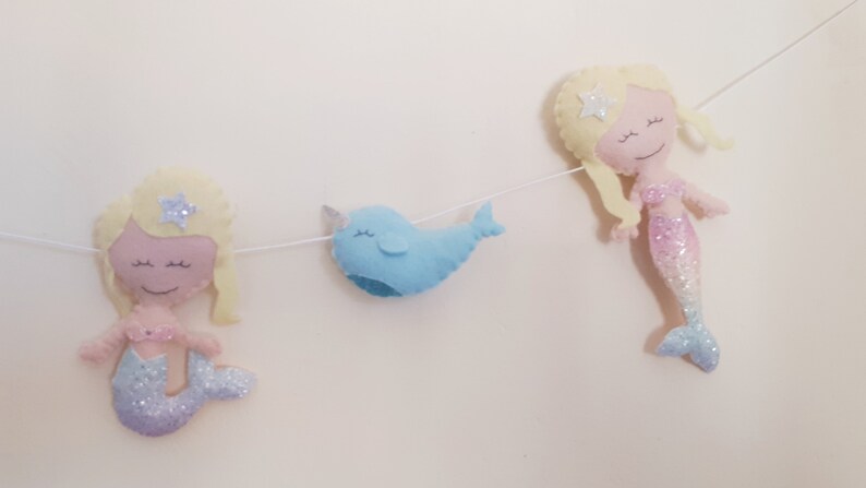 May include: A felt garland with two mermaids and a narwhal. The mermaids have blonde hair and pink and blue glitter tails. The narwhal is blue with a white horn.