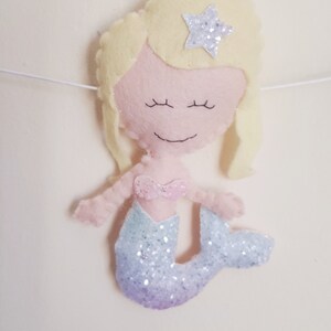 May include: A felt mermaid doll with blonde hair, a pink top, and a glittery purple and blue tail. The mermaid has a star on her head.