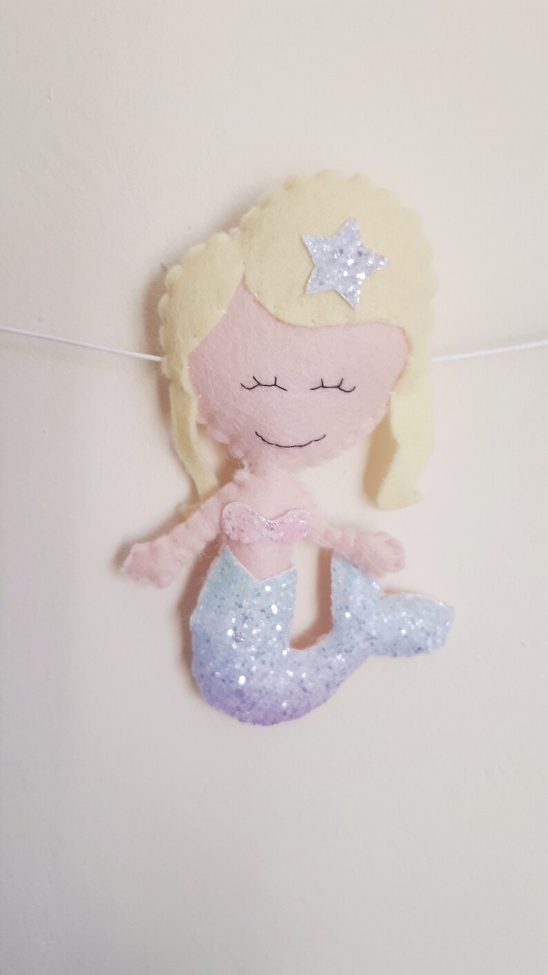 May include: A felt mermaid doll with blonde hair, a pink top, and a glittery purple and blue tail. The mermaid has a star on her head.