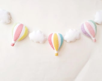 May include: A garland of pastel-coloured felt hot air balloons and clouds. The balloons are pink, yellow, and blue with white stripes. The clouds are white.
